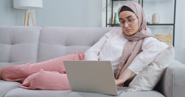 Middle plan of young muslim office worker in hijab and glasses on vacation working at a computer in home, απαντώντας σε μηνύματα ηλεκτρονικού ταχυδρομείου ενώ κάθεται σε έναν καναπέ. Μια επιχειρηματίας που δουλεύει σε καραντίνα.. — Αρχείο Βίντεο