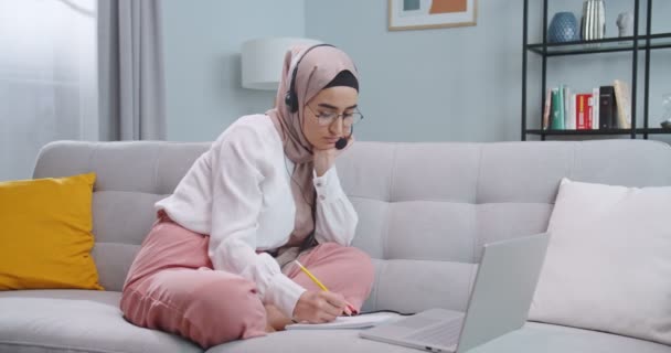 Middle plan of muslim freelancer working on laptop, μιλώντας με πελάτες που χρησιμοποιούν ακουστικά. Γυναίκα αναζήτηση για πληροφορίες, ανάγνωση ειδήσεων χρησιμοποιώντας φορητό υπολογιστή στο σπίτι. Γράφοντας σημειώσεις, διδασκαλία έννοια — Αρχείο Βίντεο
