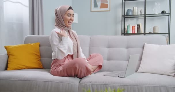 Középső terv a muszlim lány hívja barátait apartmanok segítségével egy videó link, integetett kézzel, ül egy kanapén egy stílusos szobában előtt számítógép képernyőjén videobeszélgetés segítségével laptop. — Stock videók