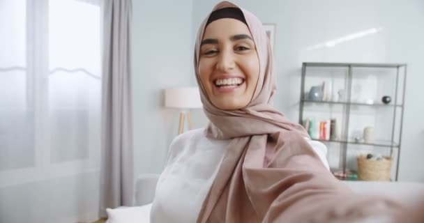 Primer plano de la joven musulmana en hijab teniendo un videochat en el fondo de la habitación moderna y hablando. En interiores. POV de la encantadora y feliz chica hablando alegremente a la cámara teniendo videochat . — Vídeos de Stock