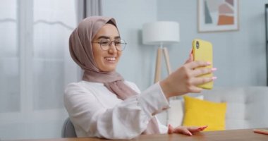 Gözlüklü gülümseyen Asyalı Müslüman kadın, ailesiyle video görüşmesi yapıyor, telefon kamerasıyla arkadaşlarıyla konuşuyor, sarı cep telefonuyla iş grubu sohbeti yapıyor. Evde çalışmak, uzaktan çalışma kavramı.