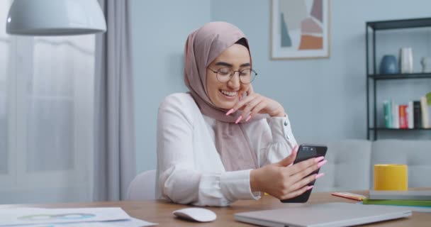 Wanita muslim Asia yang bahagia berkacamata membuat panggilan video ke keluarga, berbicara dengan teman menggunakan kamera depan telepon, melakukan obrolan kelompok bisnis. Bekerja di rumah, konsep kerja terpencil isolasi diri. — Stok Video