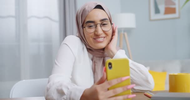 Närbild av ung muslimsk kvinna bär hijab och glasögon med videosamtal med kamera på gul smartphone medan du sitter på mysiga eleganta moderna hemmakontor. Ung asiatisk student talar med familjen. — Stockvideo