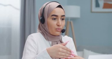 Duygusal genç Müslüman kadın satış temsilcisi. Başörtülü ve kulaklıklı kız müşteriyle konuşuyor, video konferans görüşmesi yapıyor, dizüstü bilgisayara bakıyor, web kamerasıyla konuşuyor, internet üzerinden satıyor, evden çalışıyor..