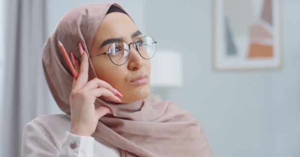 Primer plano retrato de la joven hermosa mujer musulmana en un hijab y gafas con pluma en las manos mirando soñadora pensativamente en alguna parte, sonriendo y mirando a la cámara. Chica trabajando en casa oficina . — Vídeos de Stock