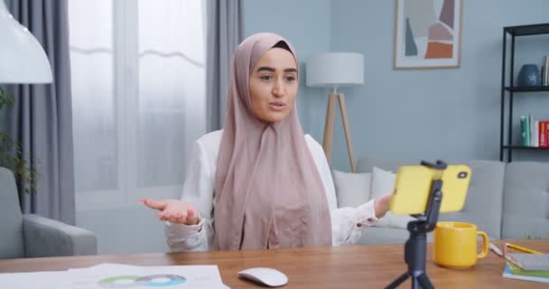 Närbild porträtt av ung attraktiv muslimsk bloggare i hijab talar på kameran skrattar glatt inomhus med mysigt modernt vardagsrum i bakgrunden. Innehåll för online vlog, sociala medier koncept. — Stockvideo