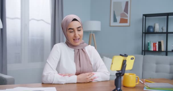 Close up do jovem blogueiro muçulmano bonito em um hijab falando sobre câmera gesticulando dentro de casa, usando smartphone amarelo. Conteúdo para vlog on-line, profissão profissão moderna trabalho, conceito de mídia social . — Vídeo de Stock