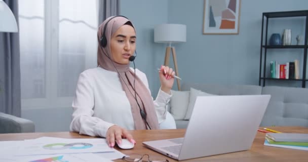 Hermoso musulmán inspirado freelancer trabajando en el ordenador portátil, hablando con los clientes con auriculares. Mujer buscar información y leer noticias en la red social utilizando ordenador portátil en el interior del hogar . — Vídeo de stock