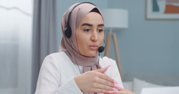 Emotionale junge muslimische Verkäuferin. Mädchen im Hijab und Kopfhörer sprechen mit Kunden, machen Videokonferenzen, schauen auf Laptop, sprechen per Webcam, verkaufen online, arbeiten von zu Hause aus. — Stockvideo