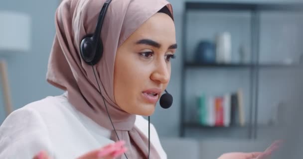 Primer plano retrato de mujer musulmana hermosa inteligente mirando en la pantalla del ordenador portátil y sentado en la oficina en casa. Mujer en el operador del centro de llamadas utilizando auriculares y micrófono que explora la información a los clientes . — Vídeo de stock