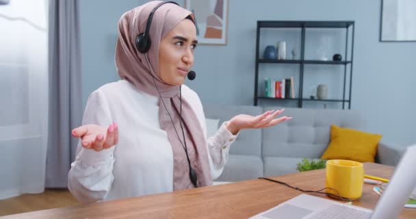 Primer plano de la joven hermosa mujer musulmana en un hijab con auriculares, videollamadas cliente distante en el ordenador portátil. Consultor que habla con el cliente usando la aplicación de computadora de chat virtual . — Vídeo de stock