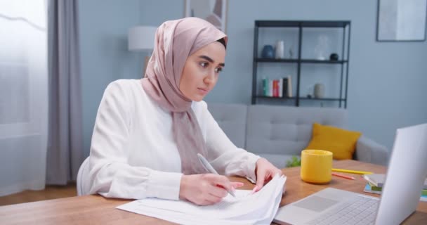 Plano del lado medio de una estudiante musulmana que estudia en el escritorio de la habitación, usando un ordenador portátil y libros, escribiendo papeles. Chica con hijab preparándose para los exámenes. Estilo de vida de los estudiantes, concepto islam moderno — Vídeos de Stock