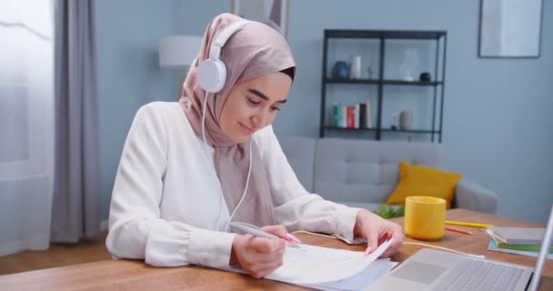 Plano médio de estudante muçulmano vestindo hijab, fones de ouvido estudando na mesa no quarto, usando laptop e livros, escrevendo papéis, ouvindo música, se preparando para exames. Estilo de vida estudantil, conceito islâmico moderno — Vídeo de Stock