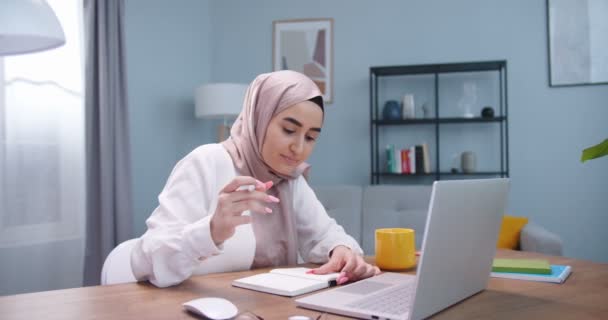 ラップトップで働いている若い学生イスラム教徒の女の子は、インターネットの先生とオンラインで勉強します。ヒジャーブの女性は、ラップトップを使用してインターネット読書ニュースで情報を検索し、自宅でノートブックにそれを書き込みます. — ストック動画