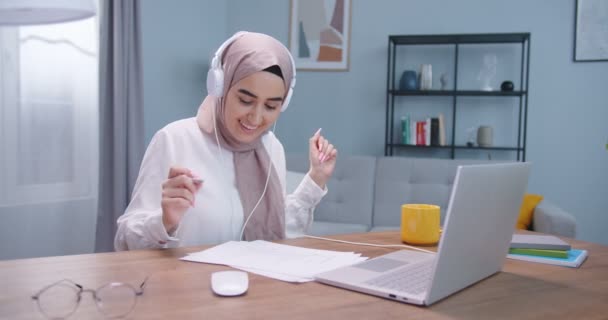 Plano medio de un hermoso estudiante musulmán que usa hijab, auriculares que estudian en el escritorio de la habitación, usando computadora portátil y libros, escuchando música y bailando mientras se prepara para los exámenes. Concepto de estilo de vida . — Vídeo de stock