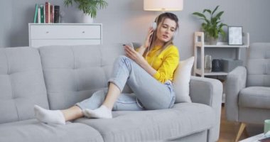 Neşeli, neşeli kız telefonundan beyaz kulaklıklarla müzik dinliyor ve kanepede dans ediyor. Vücudunu canlı ritme göre hareket ettiren bir kadın. Modern hayat kavramı, rahatlama, yaşam tarzı..