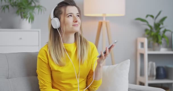 Close-up portret van een vrolijk, speels meisje luisteren muziek van haar telefoon met witte hoofdtelefoon, dansen op een levendig ritme, plezier hebben in een mooie minimalistische woonkamer. — Stockvideo