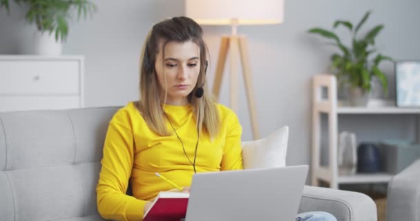 Studentinnen bearbeiten ein neues Unterrichtsthema mit einem Online-Tutor, hören dem Lehrer über Kopfhörer zu und schreiben Informationen in ein schönes rotes Notizbuch.. — Stockvideo