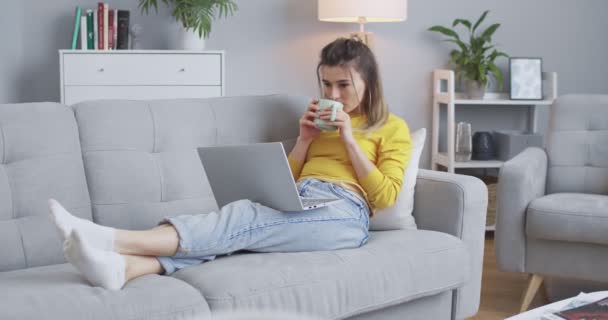 Милая девочка читает книгу онлайн, смотрит видео дома, сидя на диване в белой гостиной и держа чашку ароматного капучино. Концепция уютного дома, вечерний отдых, образ жизни — стоковое видео