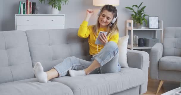 Žena si užívá hudbu z telefonu s bílými sluchátky a tancuje na gauči, baví se v krásném minimalistickém obývacím pokoji. Koncept moderního života, relaxace, životního stylu. — Stock video