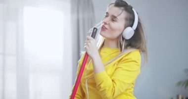 Güzel sarı bluzlu ve beyaz kulaklıklı güzel bir kızın oturma odasını temizlerken paspas ve dans kullanarak şarkı söylediği ön portreyi kapat. Eğlence anlayışı, temizlik ve kandırmaca.