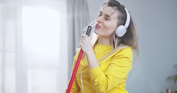 Close up front portrait of cute girl in a beautiful yellow blouse and white headsets singing using a mop and dancing while cleaning her house in living room. Concept of fun, cleaning and fooling. — Stock Video