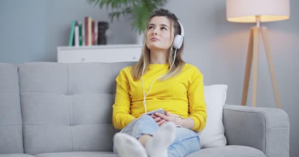 Ragazza allegra e giocosa che ascolta musica al telefono usando le cuffie, muovendo la testa in rhytm. Donna che si diverte in un bellissimo soggiorno minimalista. Concetto di auto-sviluppo, relax, stile di vita. — Video Stock