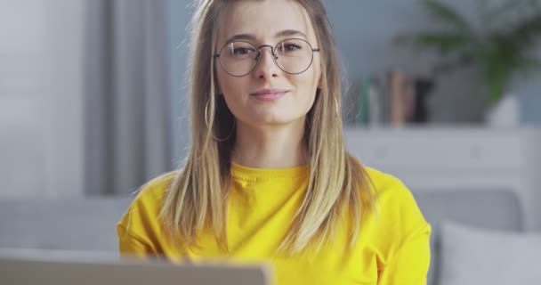 Smart blondin i gul tröja och glasögon tittar på kameran och ler. Kvinnan ser med tillförsikt fram emot med ett tillfredsställt ansiktsuttryck på stilrent vardagsrum. — Stockvideo