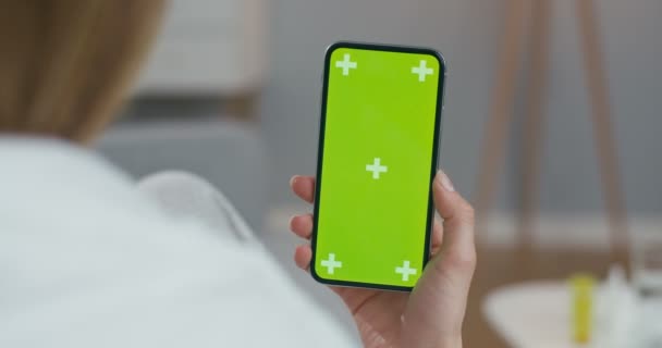 Ver por encima del hombro de la mujer en el teléfono inteligente vertical negro con pantalla verde y movimiento de seguimiento. Chica sentada en casa envuelta en manta y viendo vídeo en el teléfono celular. Clave de croma. De interior . — Vídeos de Stock