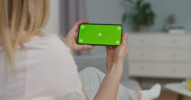 Vue au-dessus de l'épaule de la femme blonde sur smartphone horizontal noir avec écran vert et mouvement de suivi. Fille assise à la maison en quarantaine et vidéochat via webcam téléphonique. Concept de conversation par vidéochat. — Video