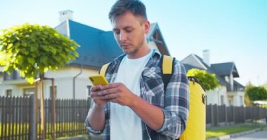 Termal sırt çantalı beyaz bir teslimatçı müşteri evini arıyor. Adresi kontrol etmek için akıllı telefonu kullanan çekici erkek kurye. Yiyecek, bakkal, paket teslimat..