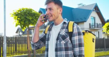 Sırt çantalı beyaz bir kurye siparişi teslim ediyor. Gülümseyen, neşeli teslimatçı akıllı telefonu kullanarak adresi kontrol ediyor, müşteriyle konuşuyor. Teslimat ve alışveriş konsepti