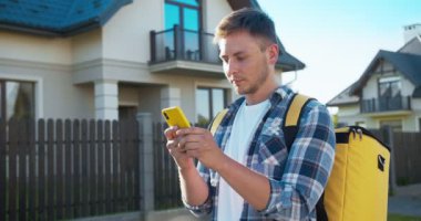 Termal sırt çantalı yetişkin beyaz kurye siparişi teslim ediyor. Yakışıklı adam akıllı telefondan müşteriye mesaj gönderiyor ve kameraya bakıp gülümsüyor. Online alışveriş ve teslimat.