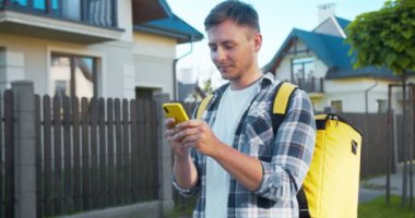 Termal sırt çantalı çekici Kafkasyalı kurye çevrimiçi siparişi teslim ediyor. Genç teslimatçı müşteriyle telefonda konuşuyor ve kameraya gülümsüyor. Teslimat servisi.