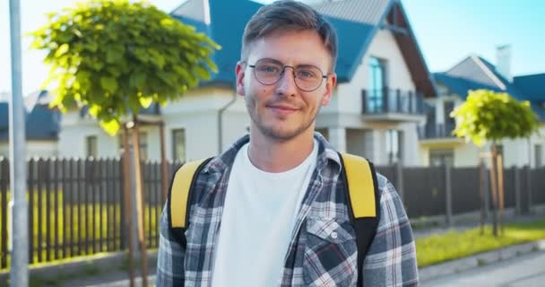 Hermoso joven mensajero masculino caucásico del servicio de entrega con gafas y una mochila térmica. Atractivo repartidor mirando a la cámara y sonriendo. Distancia social, comercio electrónico . — Vídeos de Stock