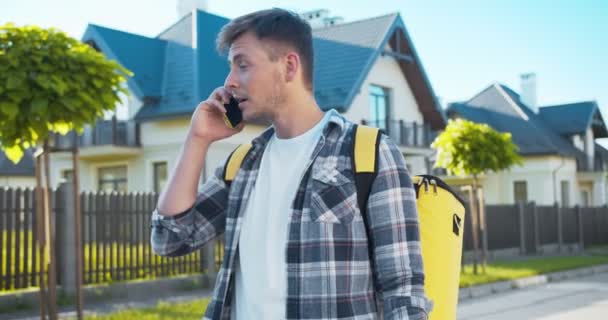 Aantrekkelijke Kaukasische koerier met een rugzak die online bestelling aflevert. Bedachtzame mannelijke werknemer van de bezorgdienst bellen en praten met de klant op zoek naar huis. Levering, shopping concept. — Stockvideo