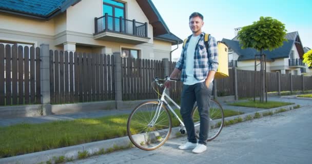 Šťastný mladý kavkazský kurýr s termálním batohem doručujícím jídlo, pyré, pošta. Hezký poslíček, stojí s bicyklem na ulici a dívá se do kamery. Online nákupní doručení. — Stock video