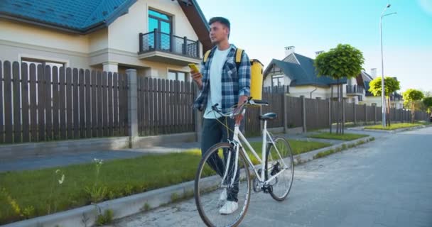 Attraktiv vit man med termisk ryggsäck från kurir tjänst leverera online beställning. Ung leverans man går ner på gatan med cykel, söker i kartor och letar efter adress. — Stockvideo