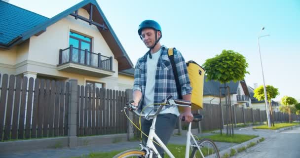 Coursier masculin adulte joyeux avec un sac à dos thermique marchant dans la rue, livrant le colis, commander. Beau livreur caucasien avec vélo regardant et frappant à la caméra. Achats en ligne. — Video