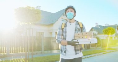 Genç beyaz erkek kurye tıbbi lastik eldiven ve maskeyle yemek getiriyor. Karton kutular taşıyan teslimatçı, içinde yiyecek, pizza ve süpermarketten aldıklarımız var. E-ticaret, online alışveriş.