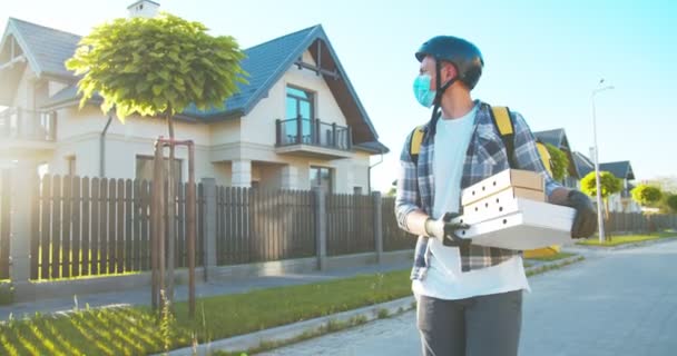 Kurir laki-laki muda Kaukasia berjalan di jalan, mengantarkan makanan dalam sarung tangan karet medis, topeng. Pengantar paket, kotak kardus dengan makanan, pizza dari supermarket. E-commerce, belanja. — Stok Video