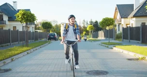 Glad vit manlig kurir rider cykeln ner till gatan leverera Internet beställning. Attraktiv ung leverans kille med en säkerhetshjälm och en termisk ryggsäck söker kunderna adress. — Stockvideo