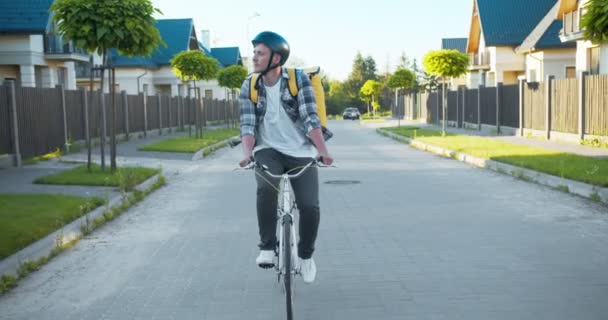 Hombre caucásico repartidor montando la bicicleta por la calle a la casa del cliente. Joven mensajero alegre con un casco y una mochila térmica entregando comida. Comercio electrónico y compras en línea . — Vídeo de stock