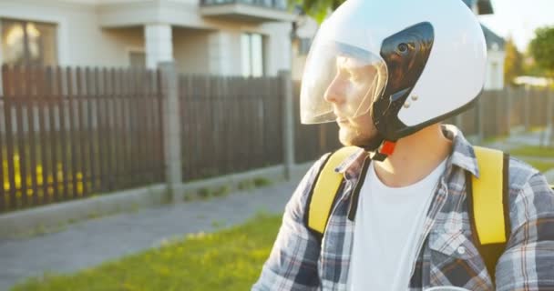 Close-up van de Kaukasische koerier in een veiligheidshelm rijden op de scooter naar de straat. Knappe jonge bezorger op zoek naar het klantenhuis. E-commerce, winkelen op internet. — Stockvideo