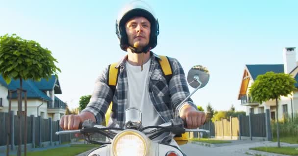 Porträt eines nachdenklichen kaukasischen Zustellers, der sich nach der Adresse des Kundenhauses umsieht. Junge männliche Kuriere fahren auf einem Motorroller auf die Straße. Essensausgabe, Selbstisolation, Quarantäne. — Stockvideo