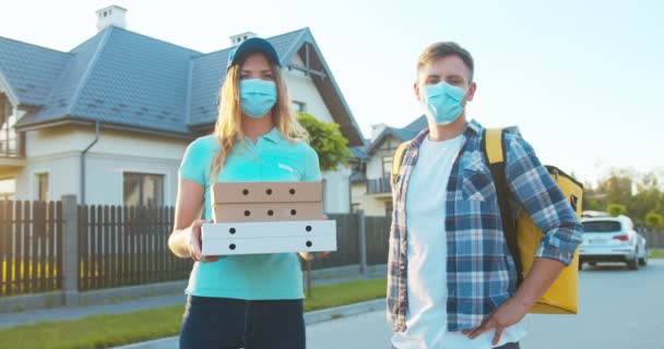 Gioiosi corrieri caucasici mascherati mascherati che consegnano ordini durante il periodo pandemico. Giovani fattorini con uno zaino e scatole di cartone che guardano la macchina fotografica. Sicurezza sanitaria. — Video Stock