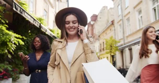 Vrolijke multi-etnische vrouwen lopen buiten met boodschappentassen en spreken op mobiele telefoons. Mooi blank meisje aan het chatten op de smartphone op straat. Afro-Amerikaanse vrouw belt op mobiele telefoon. — Stockvideo