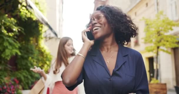 Primer plano retrato de hermosa chica afroamericana charlando en el teléfono celular en primer plano mientras camina por la calle con bolsas de compras. Mujer caucásica llamando en el teléfono inteligente en el fondo. Concepto de llamada — Vídeo de stock