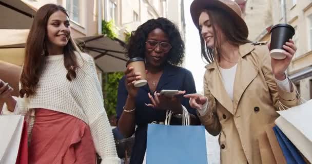 Boldog többnemzetiségű lányok sétálnak a szabadban vásárlás után. Gyönyörű afro-amerikai nő koppintott az okostelefonra, és megmutatta a barátnőinek. Kaukázusi nő egy csésze kávéval. Szórakoztató koncepció — Stock videók