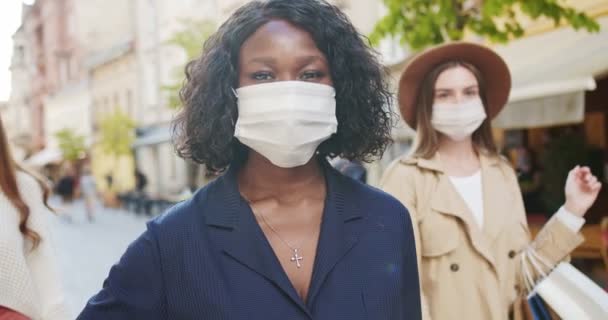 Primer plano retrato de alegre bonita mujer afroamericana en la máscara de pie al aire libre. Hermosas chicas caucásicas en el fondo con bolsas de compras en máscaras médicas en la ciudad. Concepto de cuarentena — Vídeos de Stock